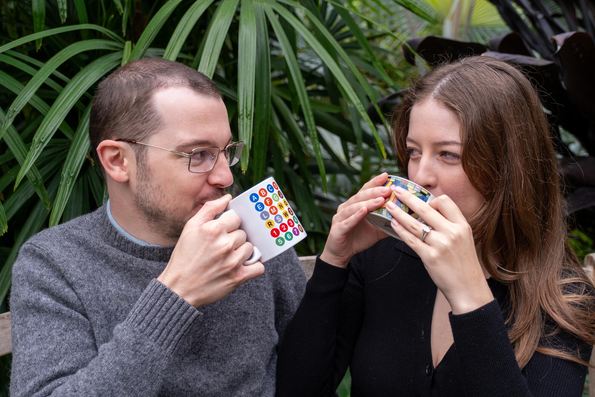 A man and a woman sip from colorful coffee mugs, looking into each other's eyes. Behind them is lush green tropical foliage.