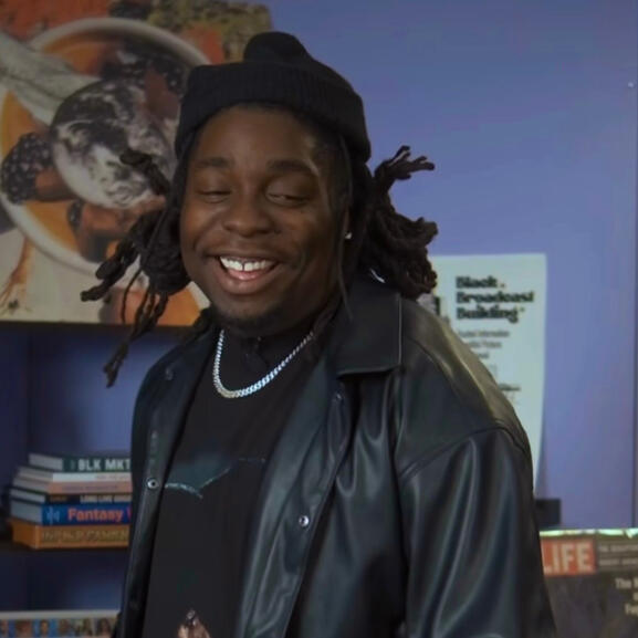A man with tied-back dreads wearing a black beanie, black leather jacket, and silver chain smiles against a purple wall.