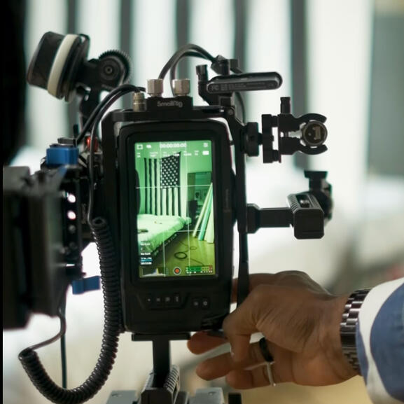 A camera with many attachments on it filming a video in vertical mode. Visible on the camera's screen is a green background with an American flag.
