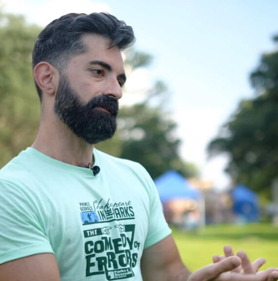 A man with short hair and a thick beard wearing a light green t-shirt in a park, talking and gesturing with his hands.