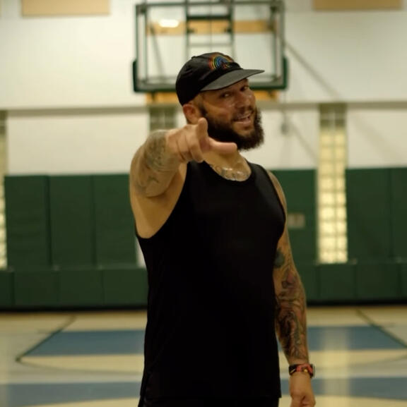 A man in a baseball cap and black tank top smiling and pointing at the camera. He is standing in a gymnasium.